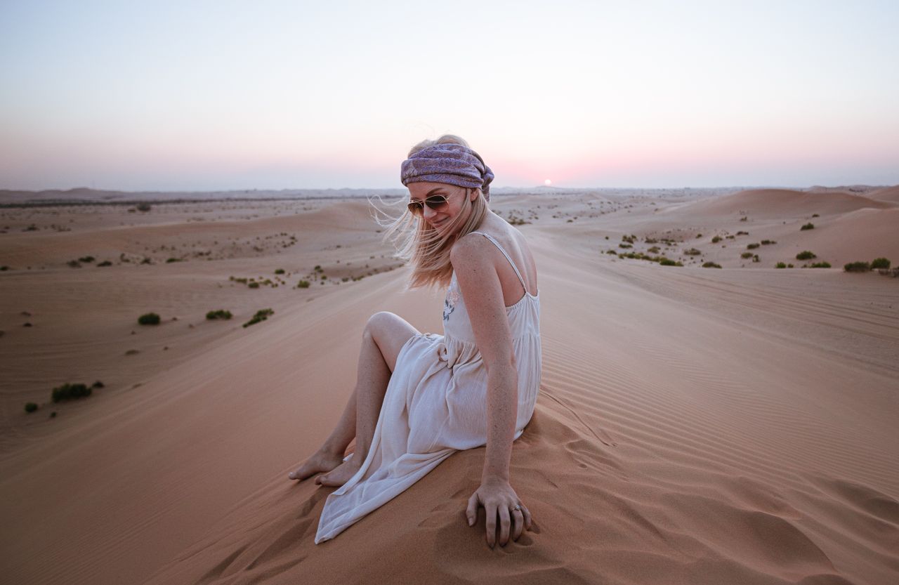 A woman in a white dress, sitting on desert sand dunes at sunset, wears sunglasses and a headscarf.